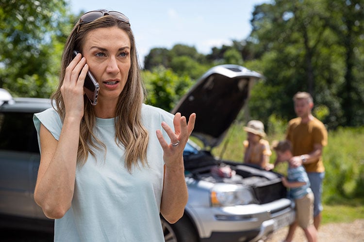 Mid adult woman who's car has broken down on a road trip calls for help with her family seen in the background near the car.